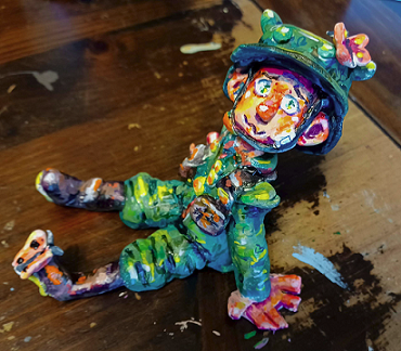 A clay sculpture of a WW1 era German soldier with butterflies on his helmet and boot. He is sitting down and staring at the camera while smiling